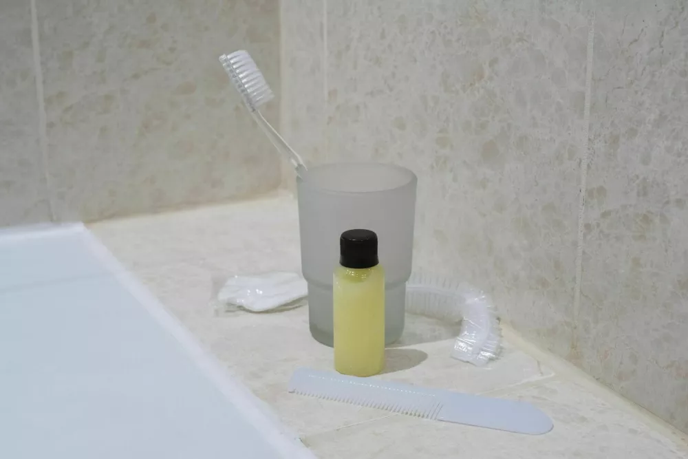 Disposable travel toiletries in hotel bathroom. Toothbrush in glass, small shampoo bottle, comb, and shower cap on marble shelf. Hospitality and travel comfort. / Foto: Irina Kononova