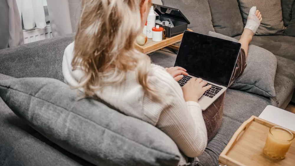 Wide angle shot of a woman resting on a comfortable sofa with her injured, bandaged leg elevated while typing on a laptop computer. On her lap tray stands a baby monitor camera, showing multitasking as a mother working from home during injury or sick leave. Modern remote work, freelancing, motherhood, health recovery, flexible workflow, and telecommuting concept. / Foto: Katharina