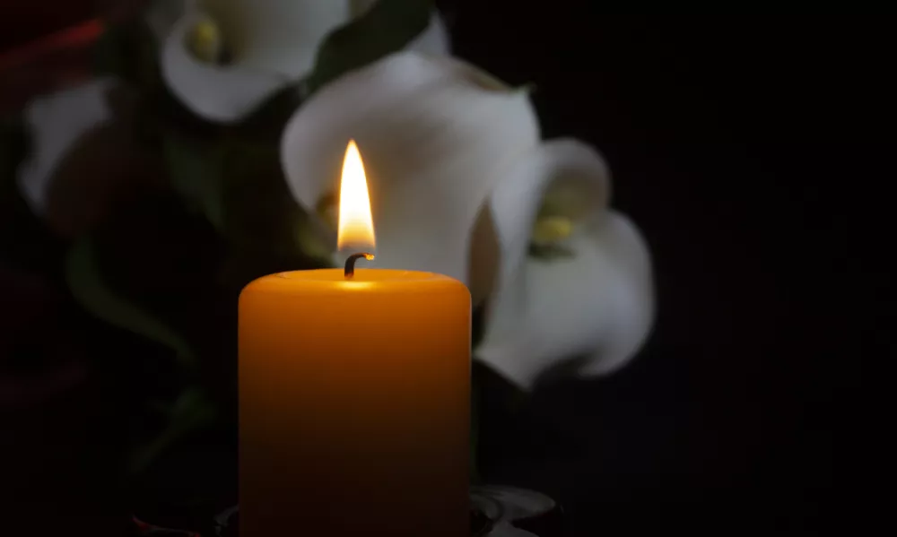 A close up of an orange candle and flame and lily flowers on a dark background.