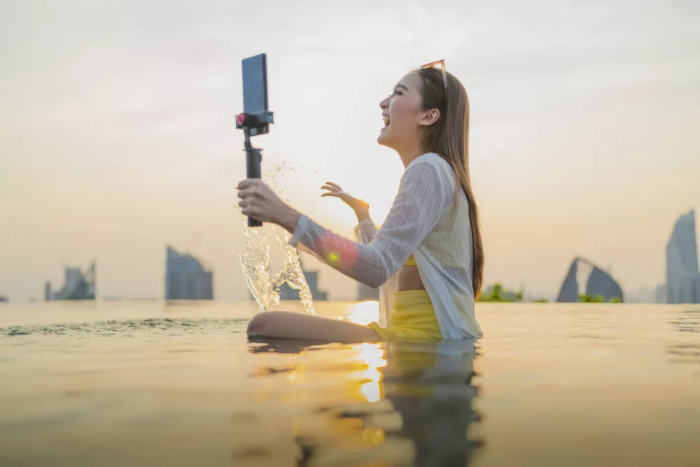 Cheerful travel influencer livestreaming vlog in rooftop infinity pool at sunset, sharing dynamic digital connection, social media engagement, joyful storytelling, wanderlust, and creator lifestyle.
