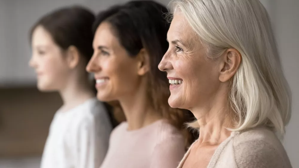 Happy attractive old 65s woman standing in row with young adult 30s daughter and little 7s granddaughter, smiling, look straight, side profile faces view. Three generations family portraits, close up